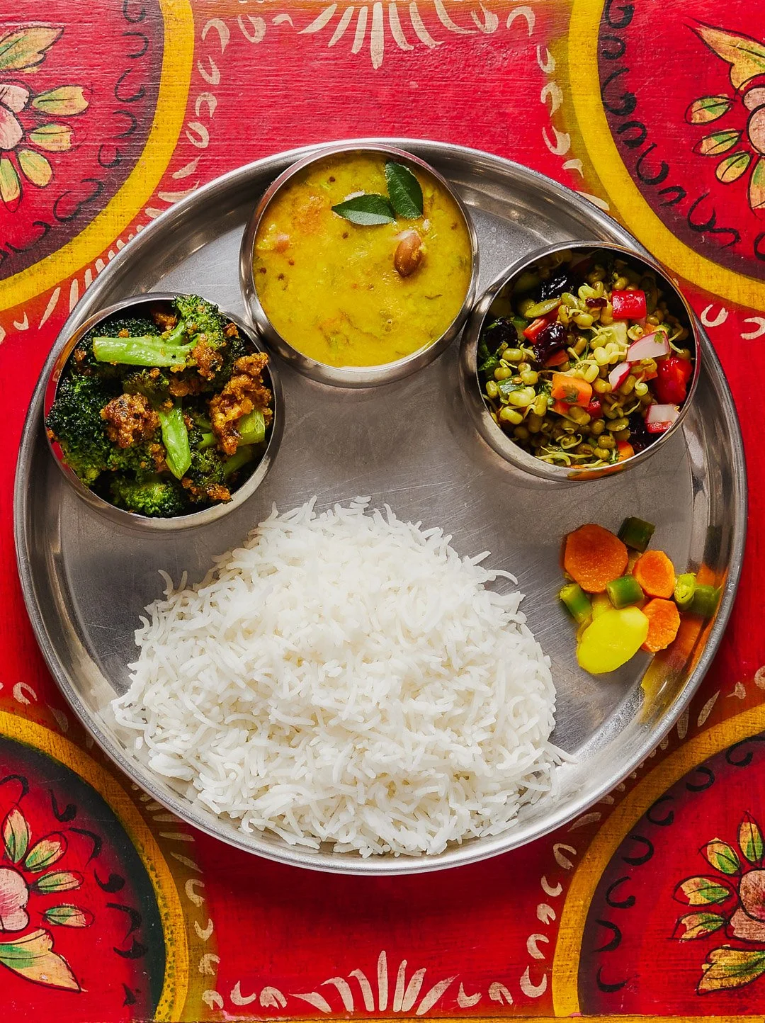 A traditional Indian meal served on a round metal platter containing white rice, vegetable curries, a mixed vegetable salad with beans and corn, cooked broccoli with spices, and pickled vegetables.