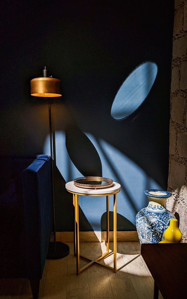 A dark room with a blue wall, a standing lamp casting shadow patterns, a small round table with a decorative tray, and a large blue and white patterned vase with a smaller yellow vase nearby.