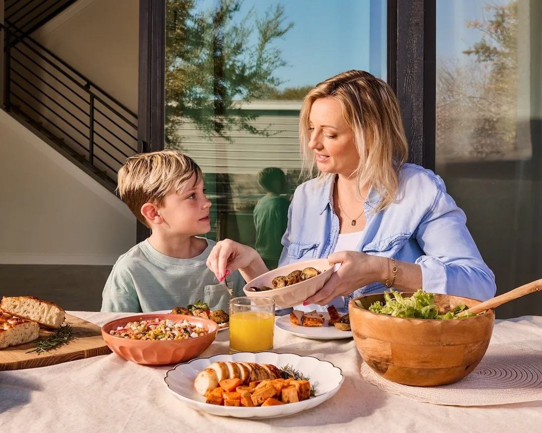 A woman and a boy sharing a meal at a table outside, with various dishes including salad, bread, roasted vegetables, and a bowl of chickpea salad.