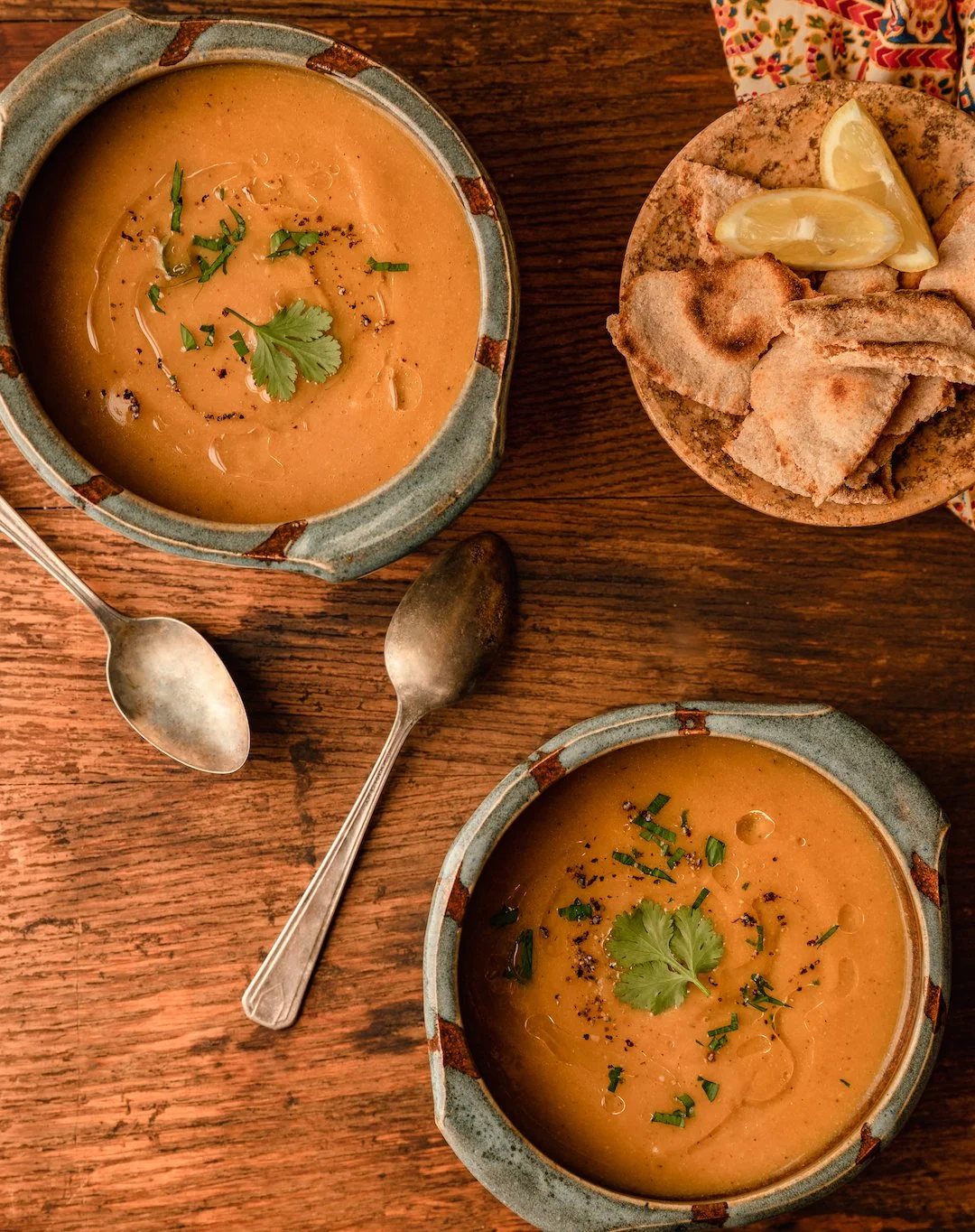Two bowls of creamy soup garnished with cilantro and chopped herbs, served with a side of fried bread slices with lemon wedges on a rustic wooden table.