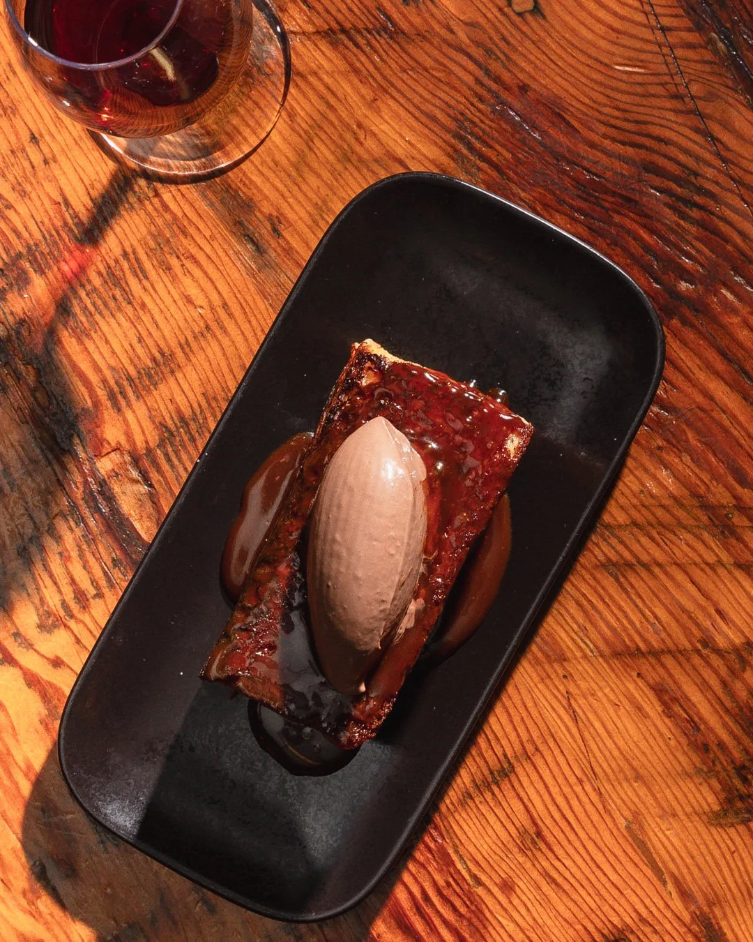 A dessert of caramelized banana with a scoop of chocolate ice cream on top, drizzled with caramel sauce, served on a rectangular black plate.