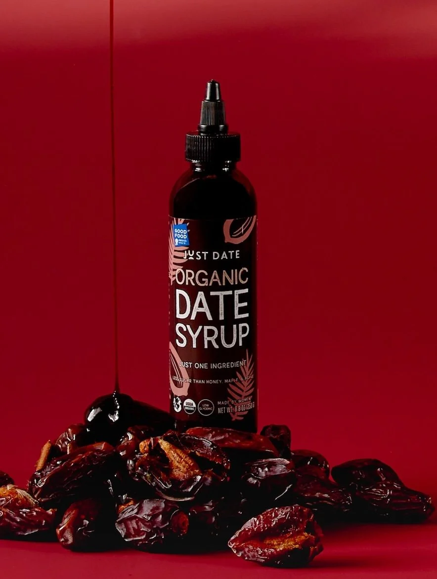 A bottle of organic date syrup with a spill of dates and date syrup on a red background.