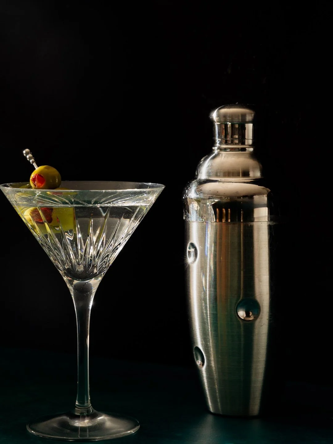 A glass martini with an olive garnish and a cocktail pick, next to a stainless steel cocktail shaker on a black background.
