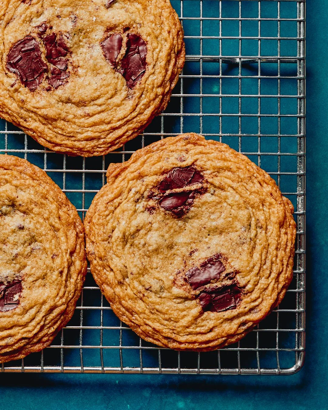 Three chocolate chip cookies on a cooling rack, with a blue surface underneath.