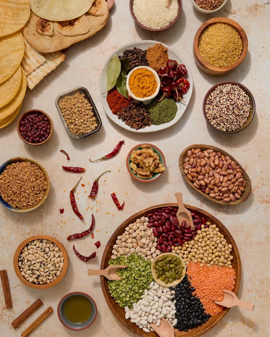Assorted spices, dried beans, lentils, and grains arranged on a table, along with some bread and dried chili peppers, used for cooking or preparing traditional Mexican dishes.