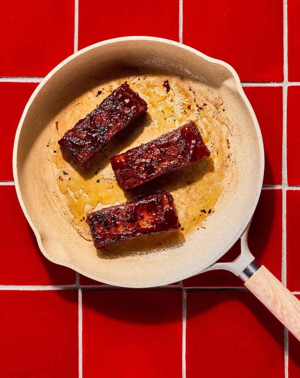 Three glazed pork ribs in a beige skillet on a red tiled surface.