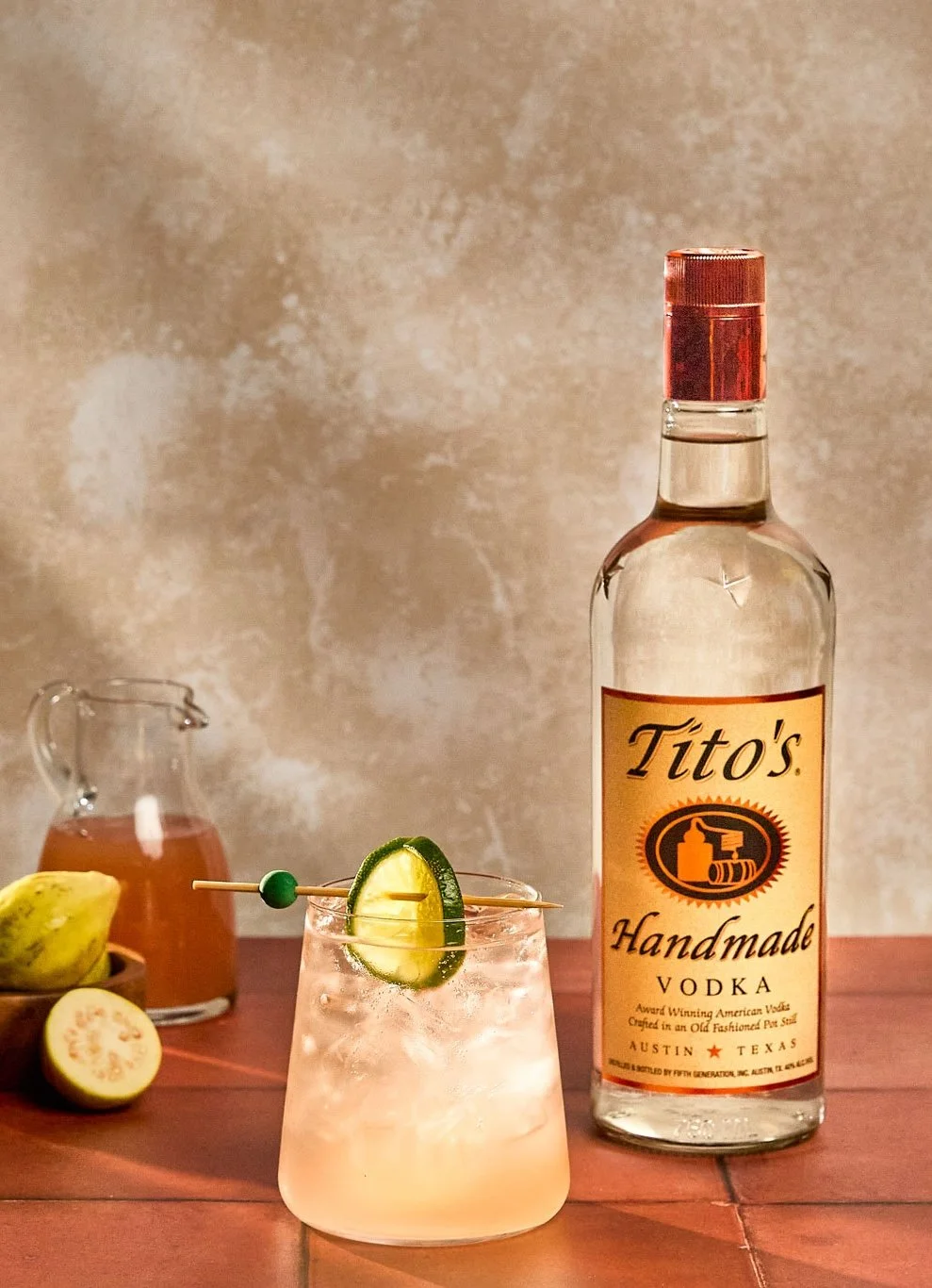 A bottle of Tito's Handmade Vodka, a glass with a lime wedge garnished drink, a lemon, and a small pitcher with a liquid, all on a wooden surface.
