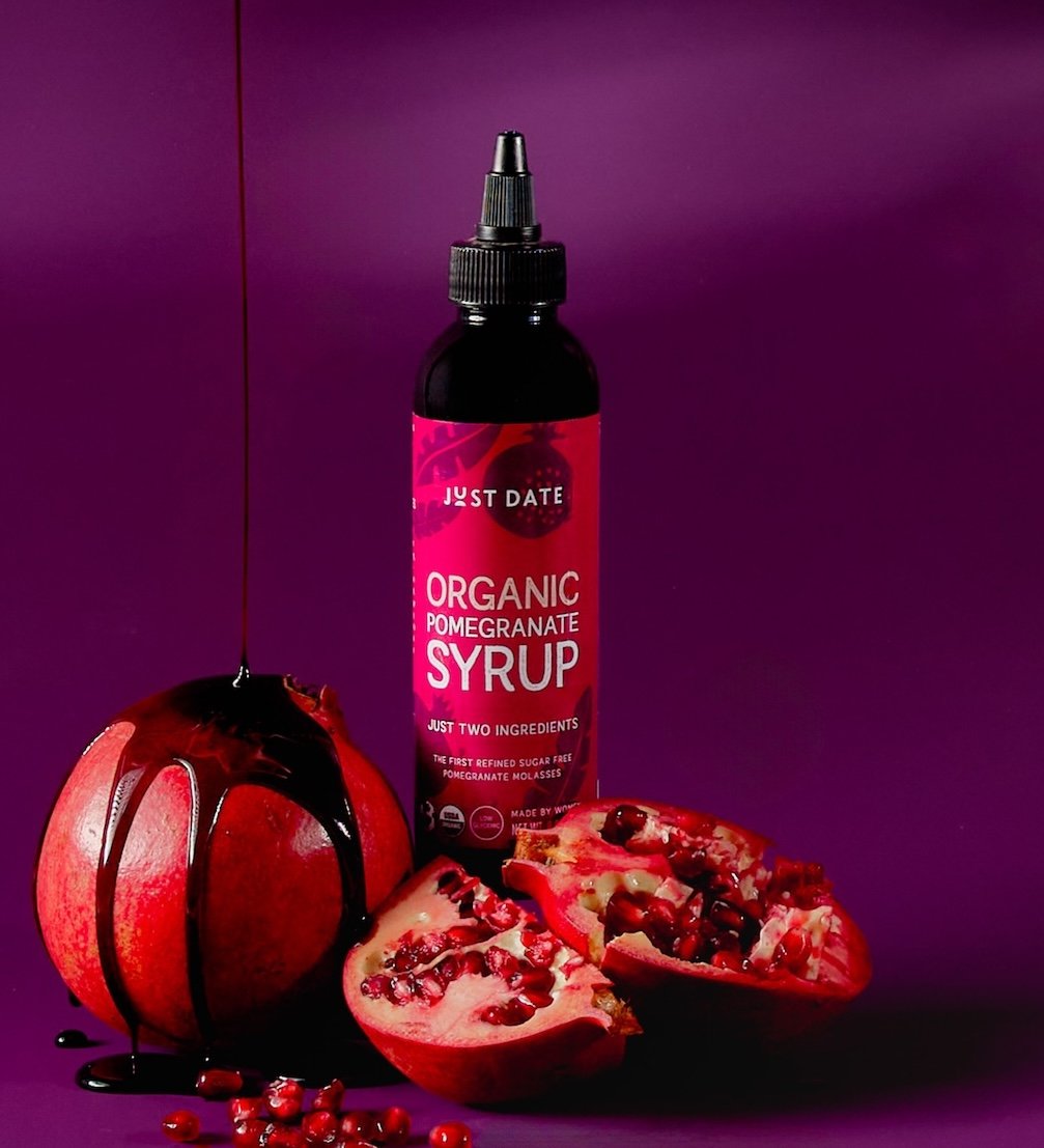 Bottle of organic pomegranate syrup, fresh pomegranate with seeds spilling out, and chocolate syrup drizzle on the fruit, with a purple background.