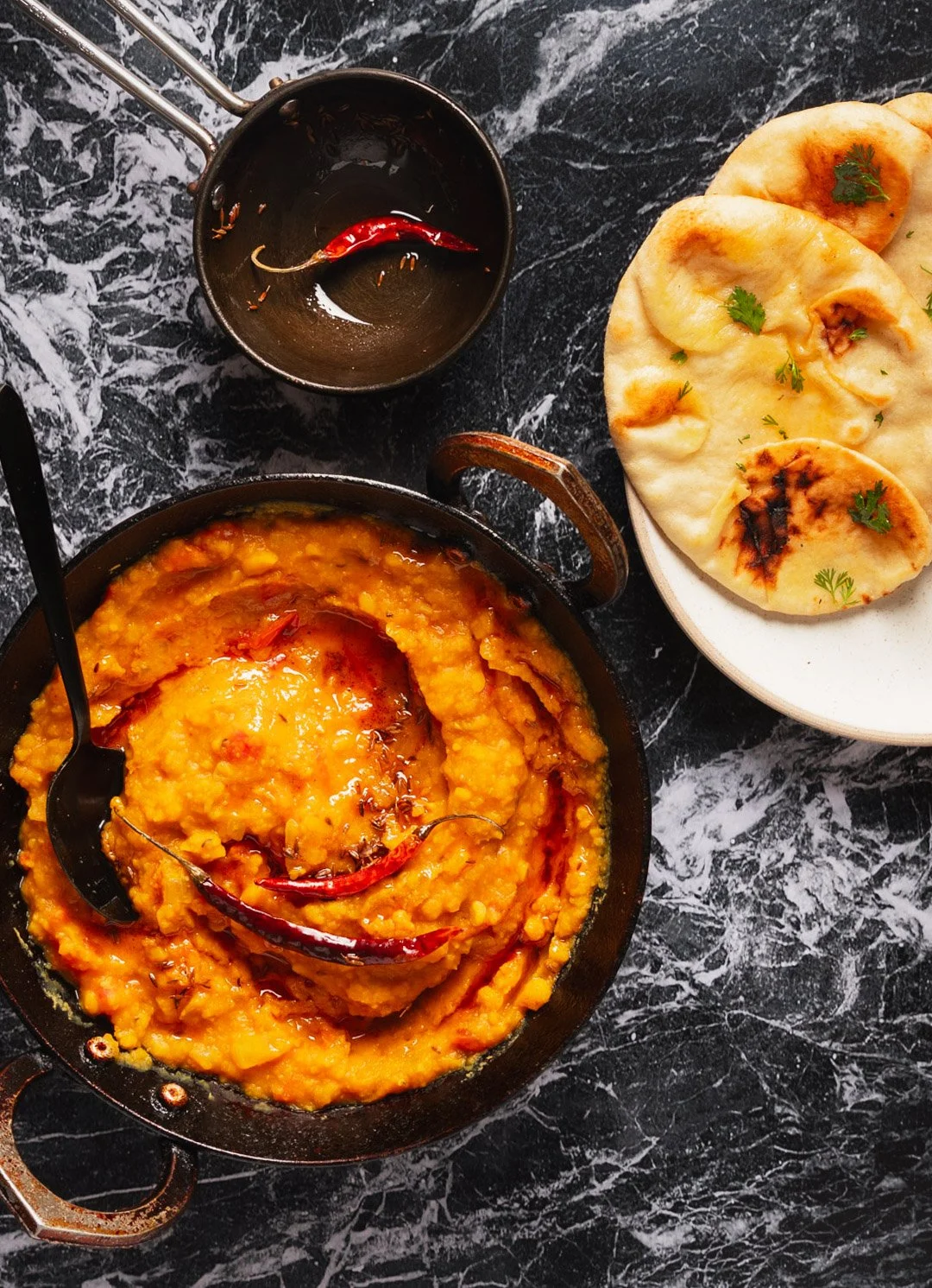 A skillet of mashed lentils topped with red chili peppers, a small black bowl with a red chili and spices, and a plate of naan bread garnished with cilantro, all on a black marble surface.