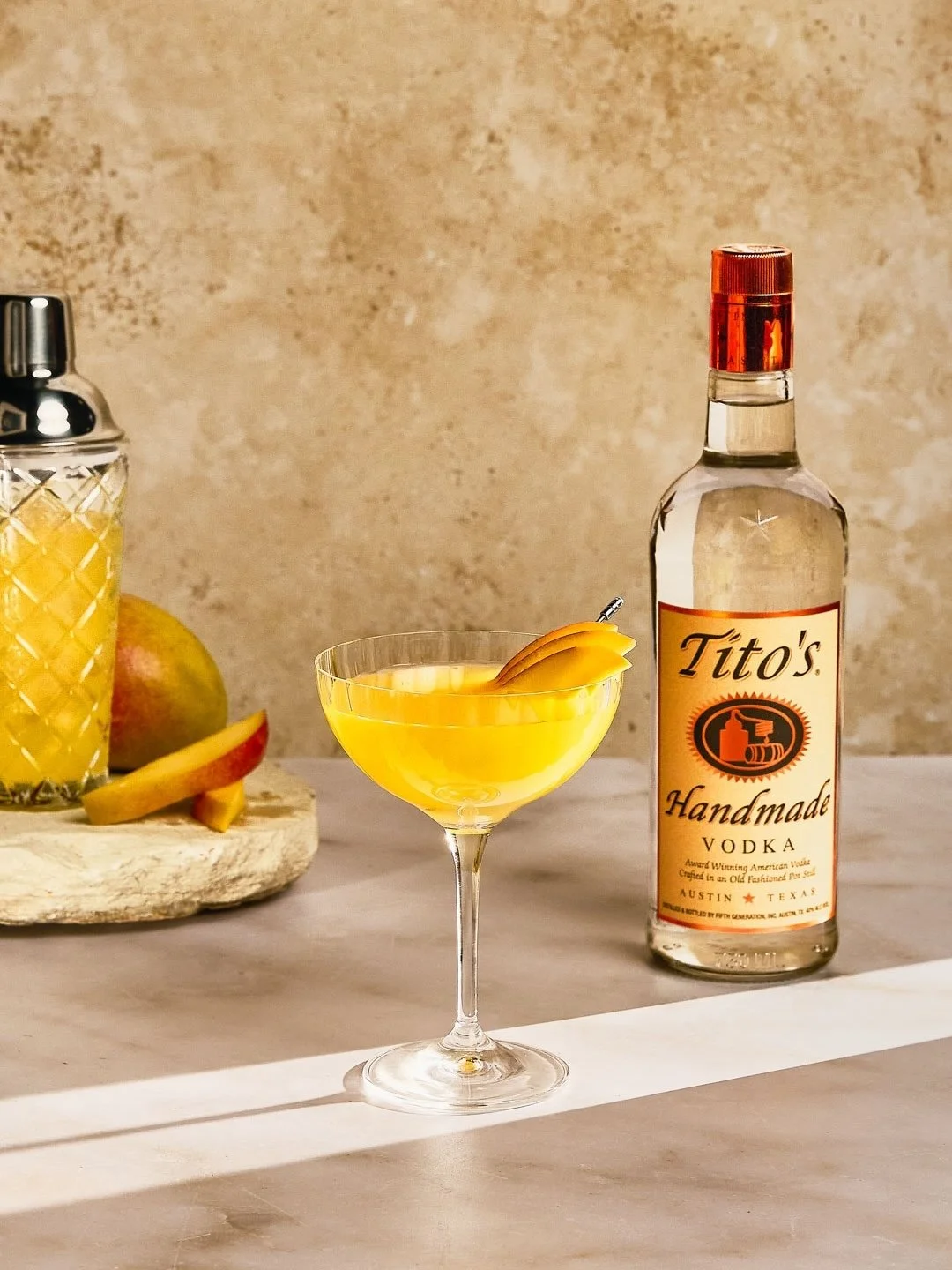 A cocktail in a coupe glass garnished with a lemon peel, a bottle of Tito's Handmade Vodka, a pineapple, and sliced fruit on a wooden cutting board on a light-colored surface against a stone wall.