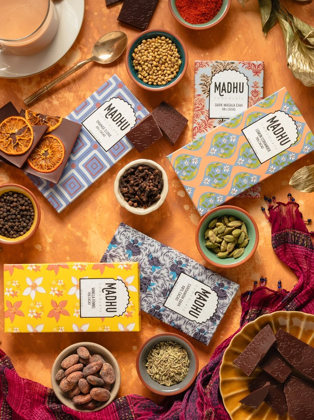 Various flavors of Madhu chocolate bars, decorative bowls of spices and ingredients, and a glass of tea or coffee on an orange table.
