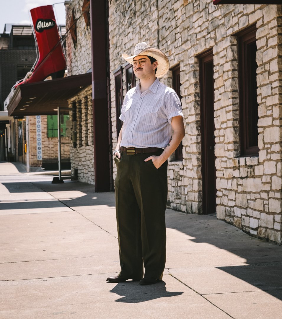 A man standing on a sidewalk next to a stone building with windows, wearing a wide-brimmed hat, glasses, a striped short-sleeve shirt, and dark pants, with his hands in his pockets and shadow cast on the ground.