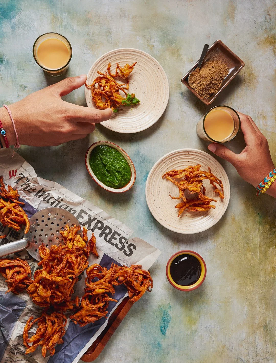 Top-down view of an Indian meal with fried onion fritters, green chutney, soy sauce, tea, and a snack, on a colorful table.