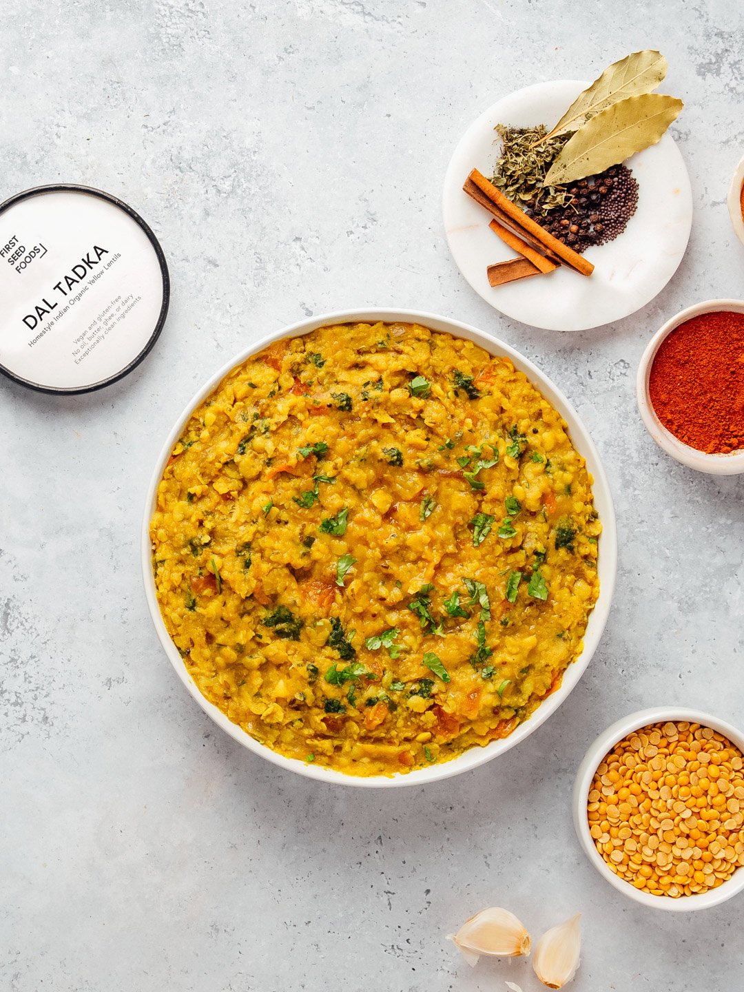 A bowl of cooked yellow lentil dal garnished with chopped herbs, surrounded by bowls of spices, garlic, and dried herbs on a light surface.