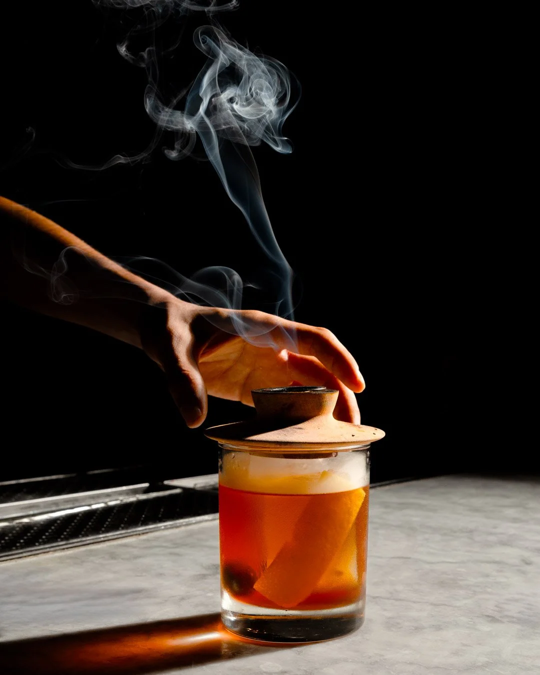 A person’s hand lifting a ceramic lid off a glass cup containing tea, with steam rising against a dark background.