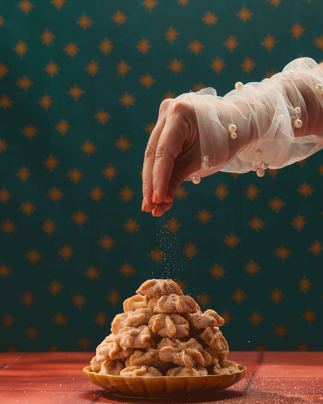 A person's hand sprinkling sugar onto a stack of breaded chicken pieces on a brown plate, with a green background decorated with orange snowflakes.