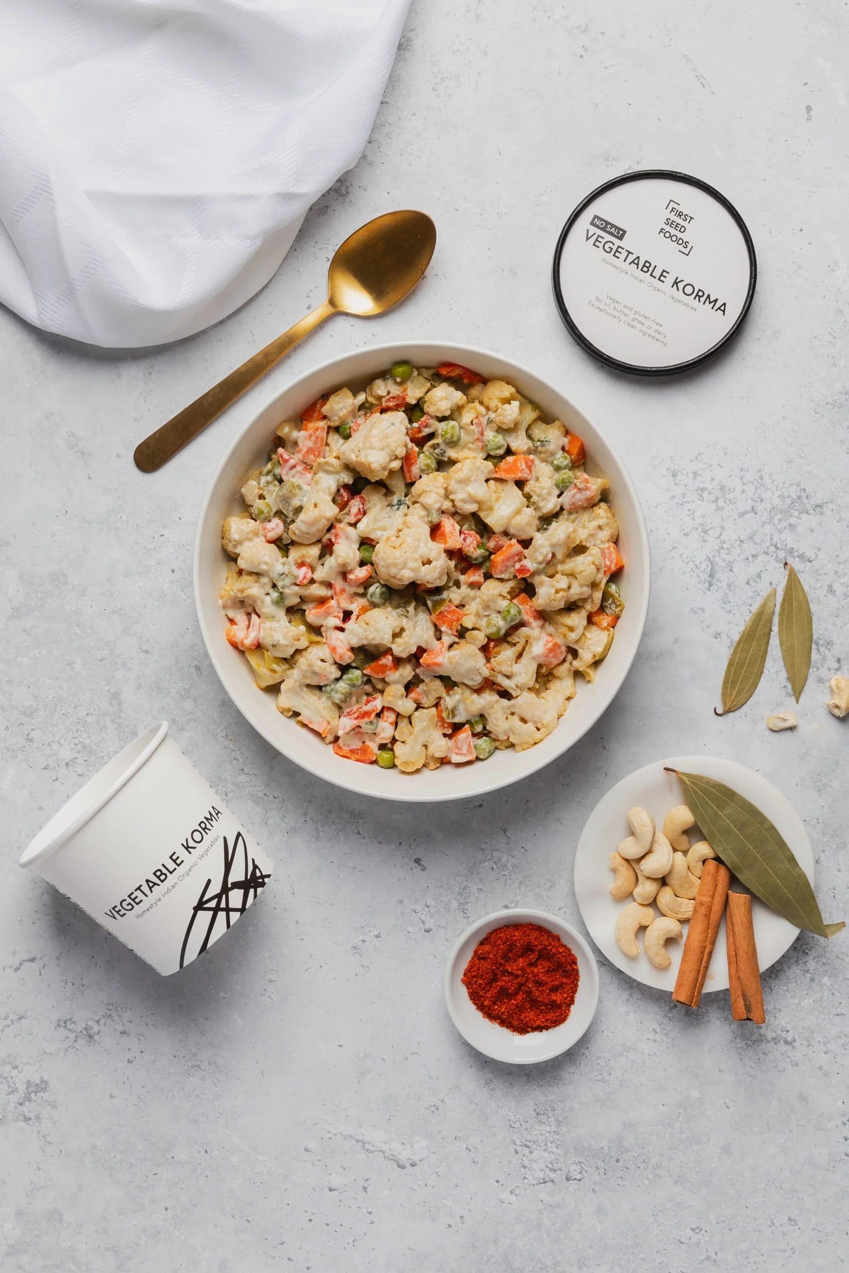 A bowl of vegetable korma with mixed vegetables in a creamy sauce, surrounded by spices, nuts, and a cup of vegan yogurt labeled 'Vegetable Korma' on a white surface.