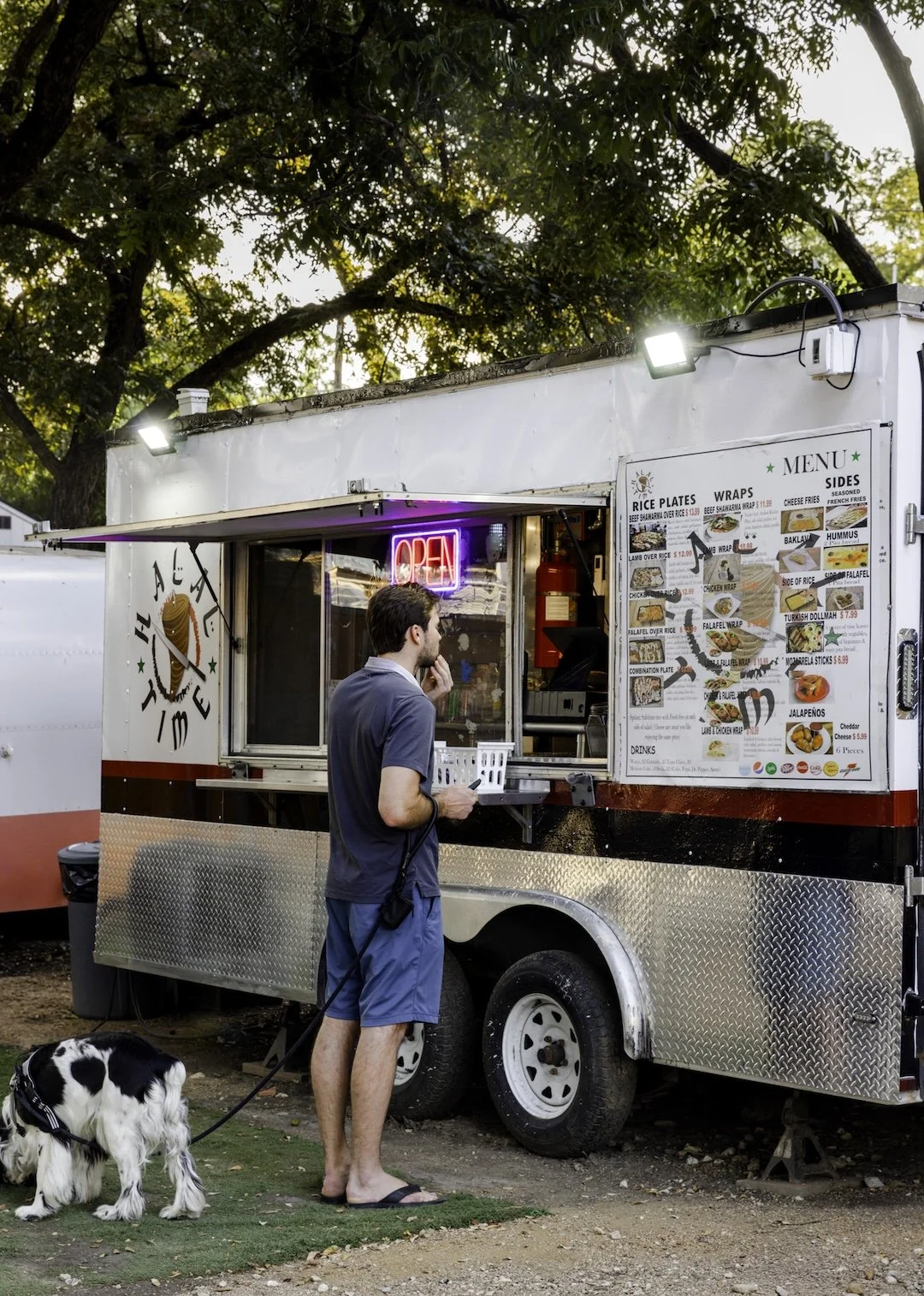 A man wearing a gray t-shirt, blue shorts, and flip-flops, holding a leash attached to a black and white dog, is ordering food at a food truck with a menu displayed outside. The truck has a neon 'OPEN' sign and a large menu showing various food optio