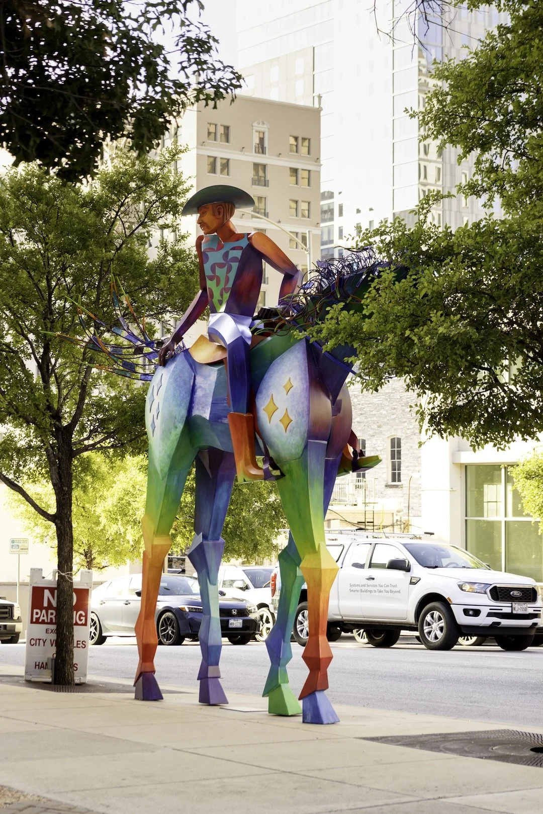 Colorful metal sculpture of a person sitting on an animal with rainbow colors, set in an urban environment with cars, trees, and buildings in the background.