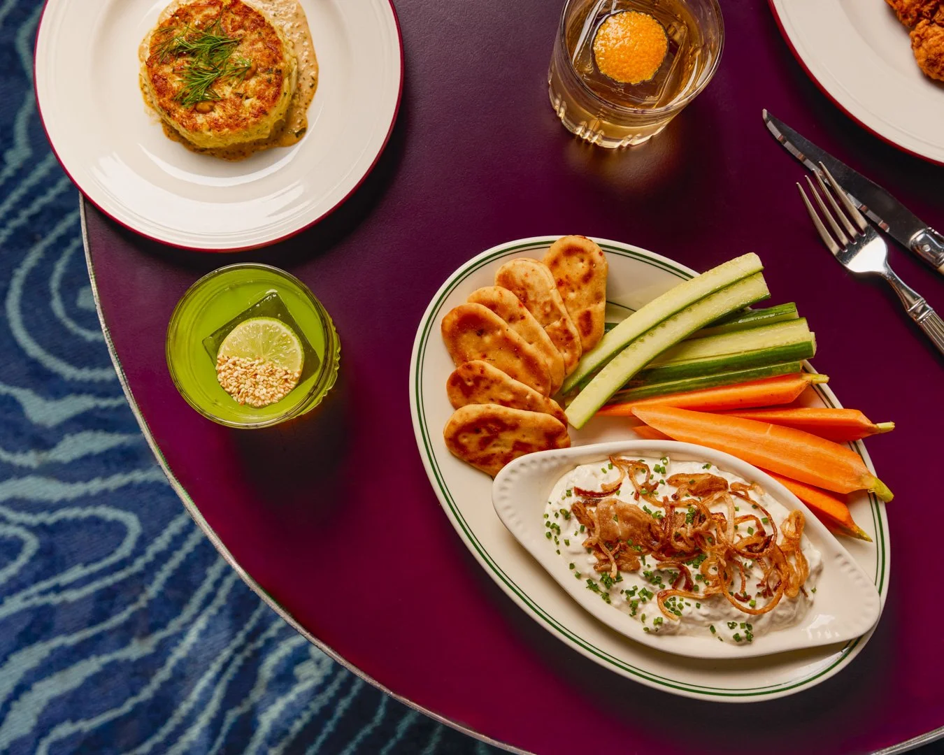 A dinner table with a plate of grilled vegetables, crackers, and dip, a bowl of sliced vegetables, and a drink with a lemon slice and sesame seeds, set on a maroon table.