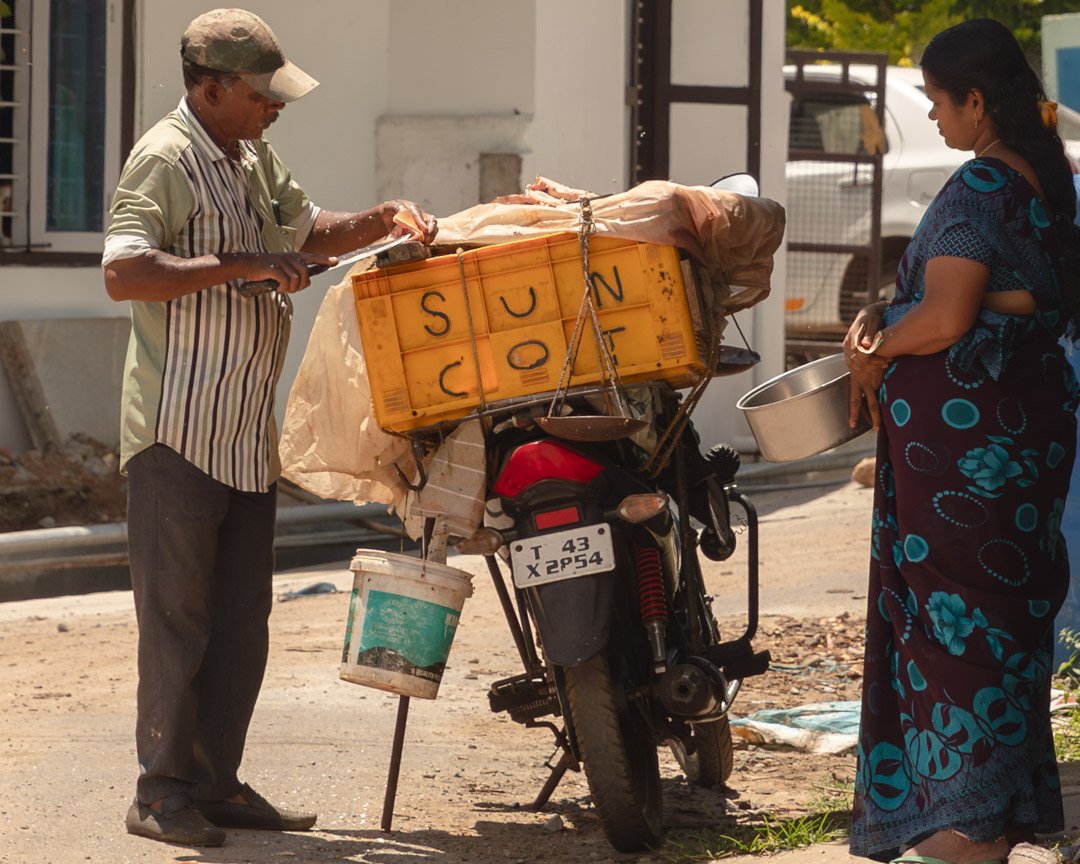 A man selling food on a motorcycle with a woman holding a metal bowl on a street.