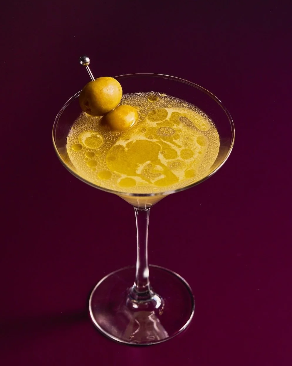A cocktail in a martini glass garnished with green olives on a pick, set against a dark purple background.