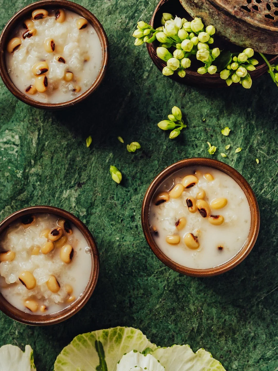 Three glasses filled with white drink topped with roasted pine nuts, on a green textured surface. There are yellow flowers and green leaves nearby.