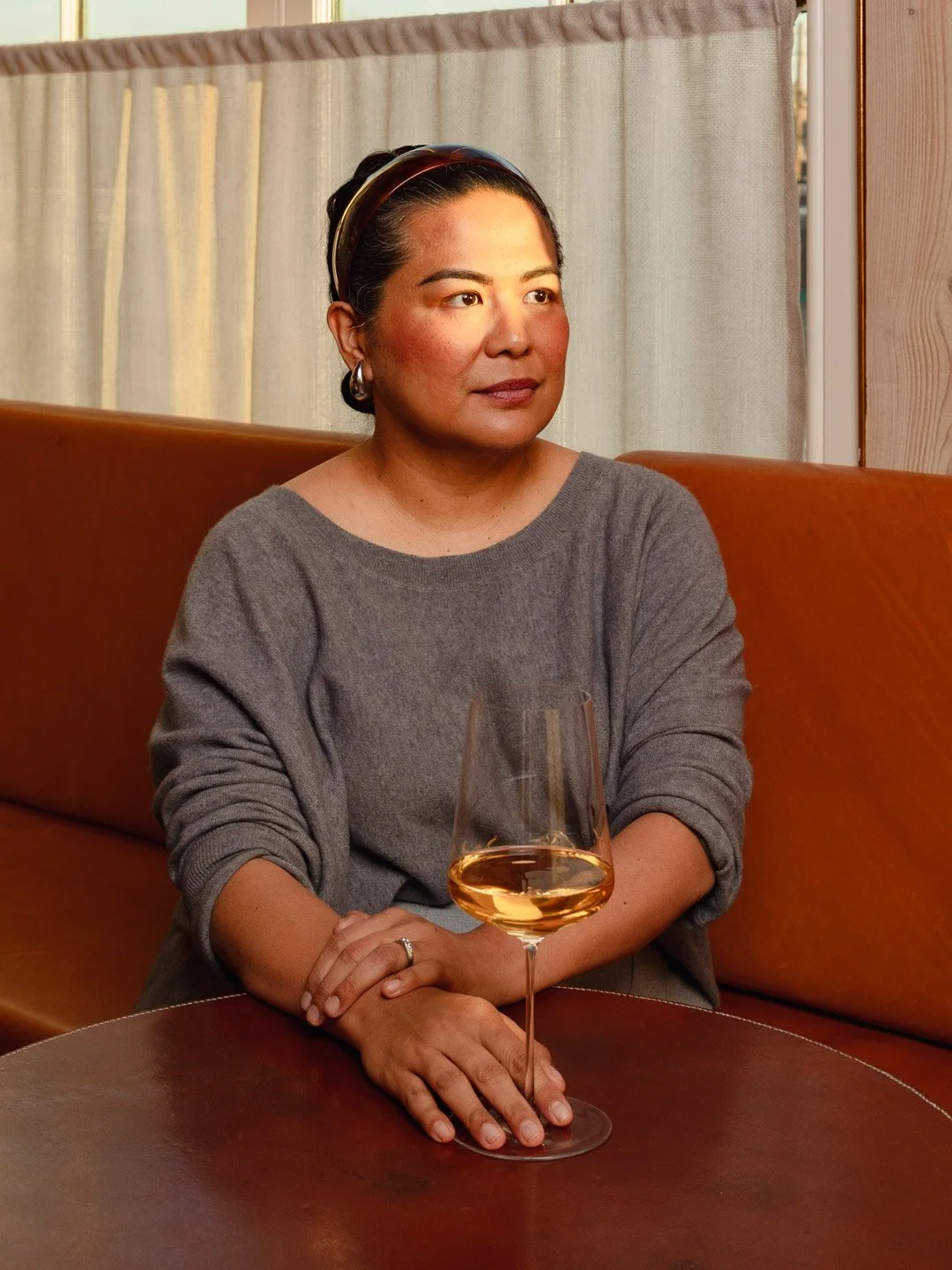 A woman with dark hair and hoop earrings sitting at a table with a glass of white wine in front of her.
