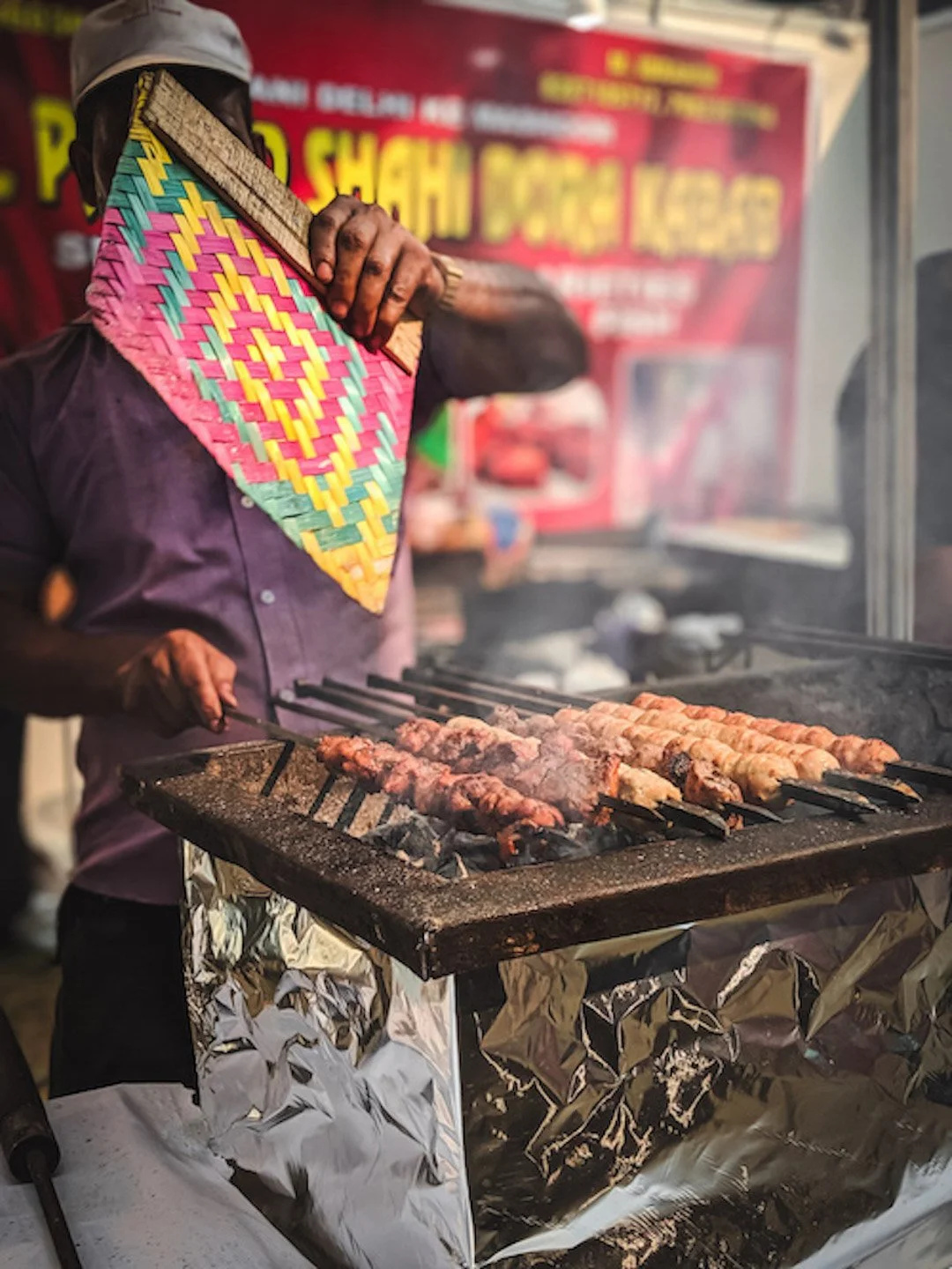 A person grilling skewered meat over an open flame at a street food stall, wearing a colorful woven scarf, a dark shirt, and a cap, with a red and yellow sign in the background.