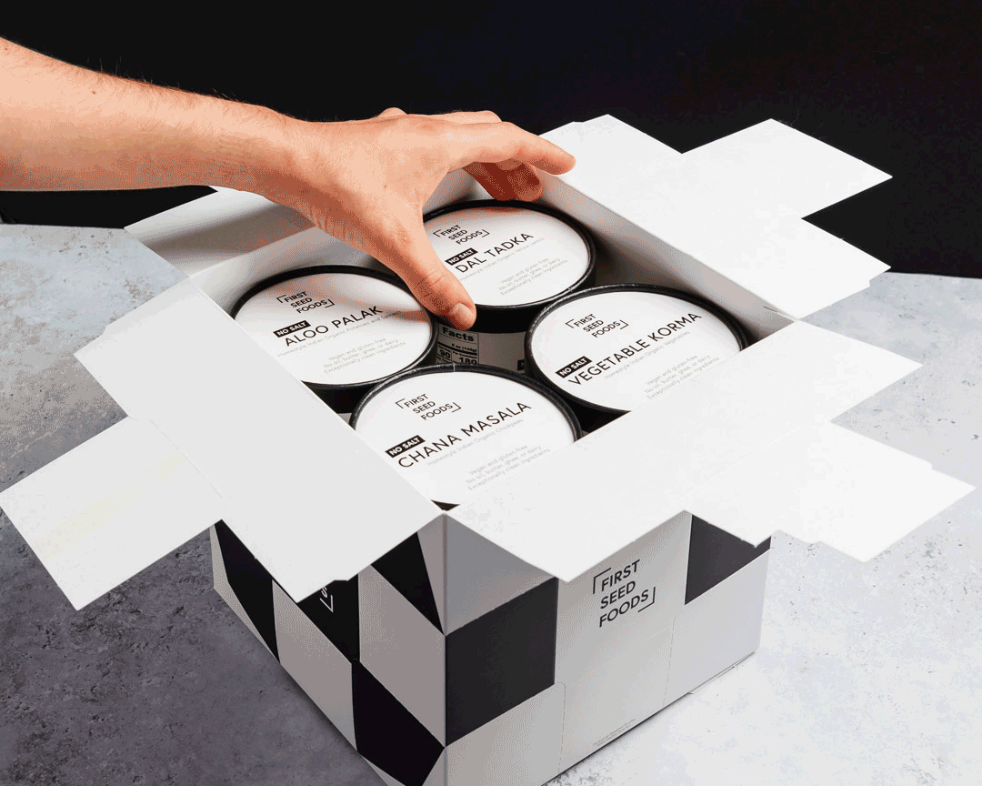 Hand opening a box of labeled food cans with a black and white geometric design, titled 'First Seed Foods.'
