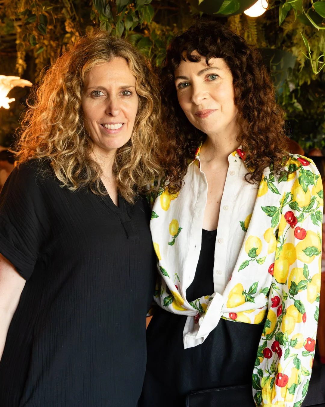 Two women with curly hair smiling, one with blonde hair wearing a black shirt, and the other with dark brown hair wearing a white shirt with a lemon and cherry pattern, in a setting with green foliage and warm lighting.