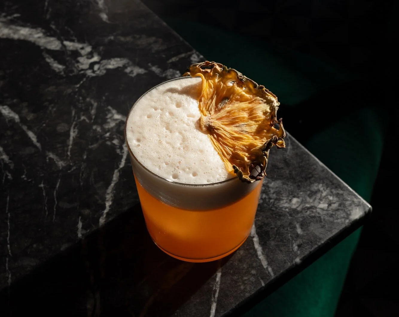 A glass of orange-colored cocktail topped with foam and a dried pineapple slice garnish, placed on a dark marble surface.