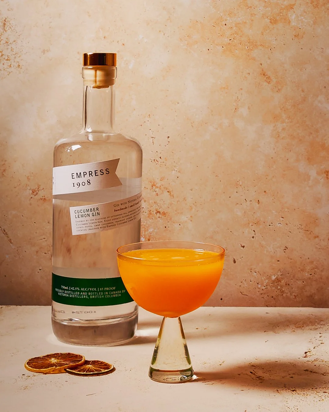 A bottle of Empress 1908 cucumber lemon gin, a coupe glass filled with an orange cocktail, and three dried orange slices on a light surface with a textured beige background.