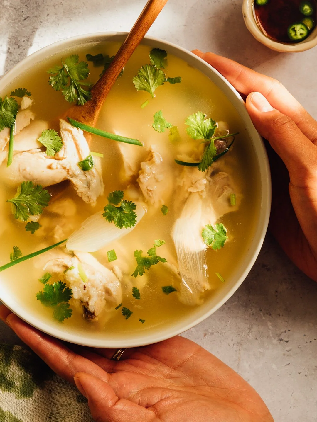 A bowl of chicken soup garnished with cilantro, green onions, and onion slices. A wooden spoon rests inside the soup. A small bowl of soy sauce with chopped green onions is visible in the background.