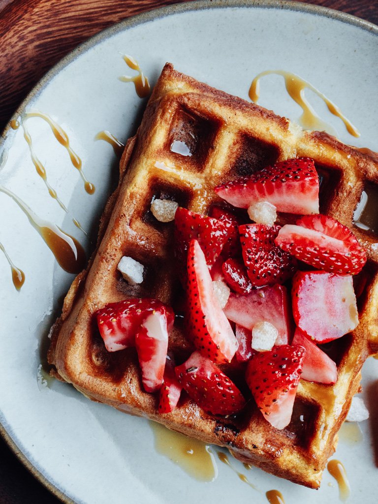 A waffle topped with sliced strawberries, small sugar cubes, and drizzled with syrup on a white plate with a drizzle of syrup on the side.
