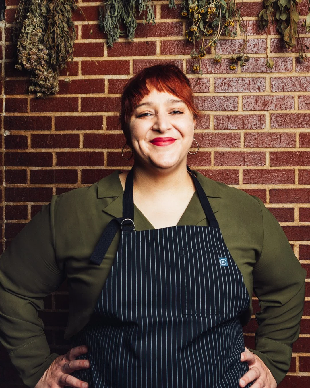 A woman with short red hair, wearing a green shirt and black apron with white pinstripes, stands in front of a brick wall with dried herbs hanging above her.