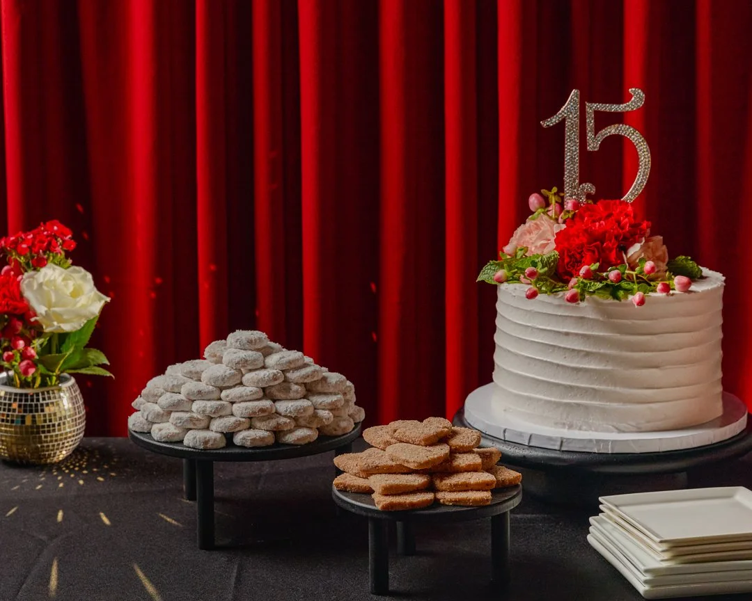 A birthday celebration table with a white frosted cake decorated with pink and red flowers and a '15' topper, accompanied by plates and a flower arrangement with red and white flowers, set against a red curtain backdrop.