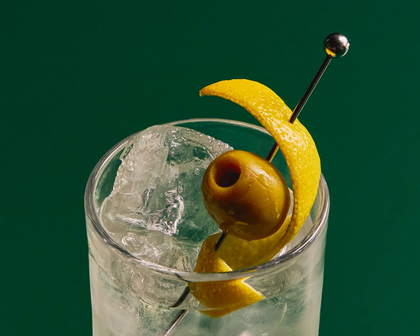 A close-up of a cocktail glass filled with ice, garnished with a lemon twist and a green olive on a cocktail pick, against a dark green background.