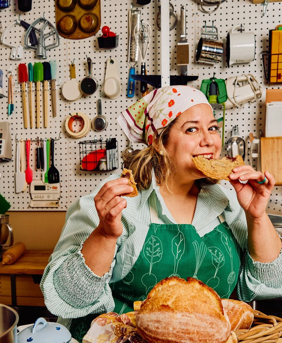 Woman wearing a green apron and a floral headscarf eats a slice of bread in a kitchen with a pegboard wall filled with kitchen tools.