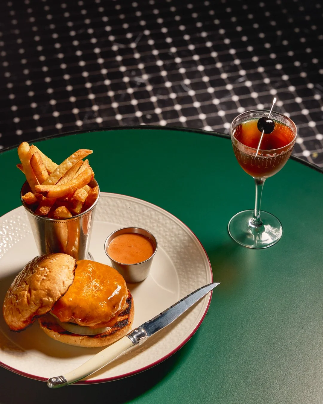 A cheeseburger with a side of French fries, a small cup of dipping sauce, and a cocktail with an olive and cherry on a green table.