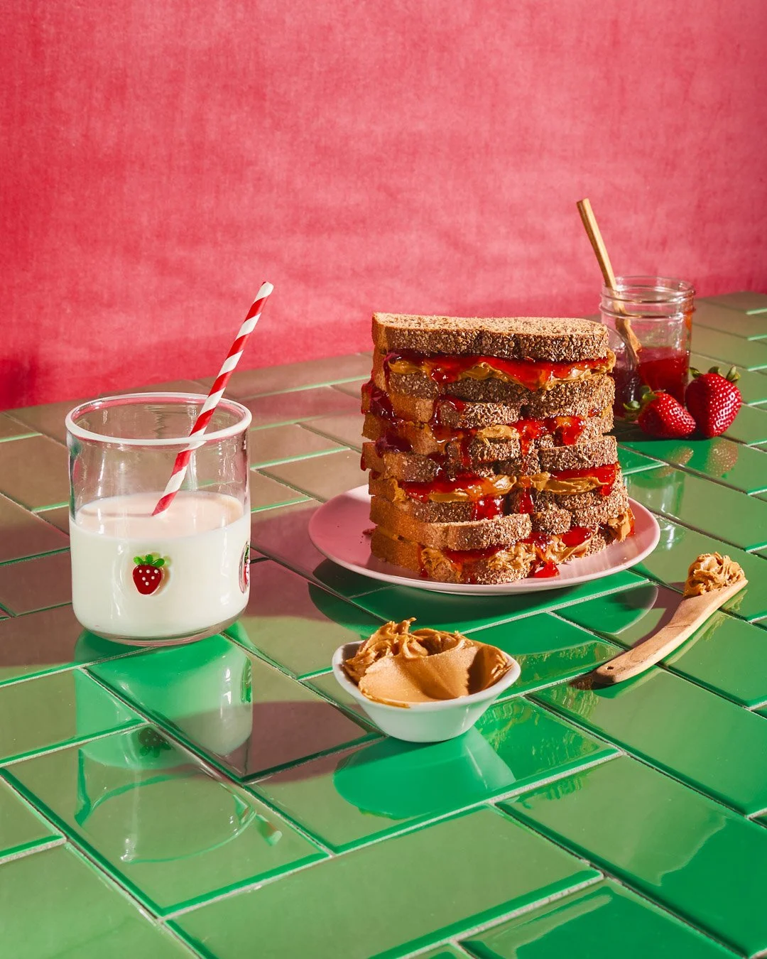 Stack of peanut butter and jelly sandwiches with strawberry jelly on green tiled table, beside a glass of milk, strawberries, and jars of jelly and peanut butter.