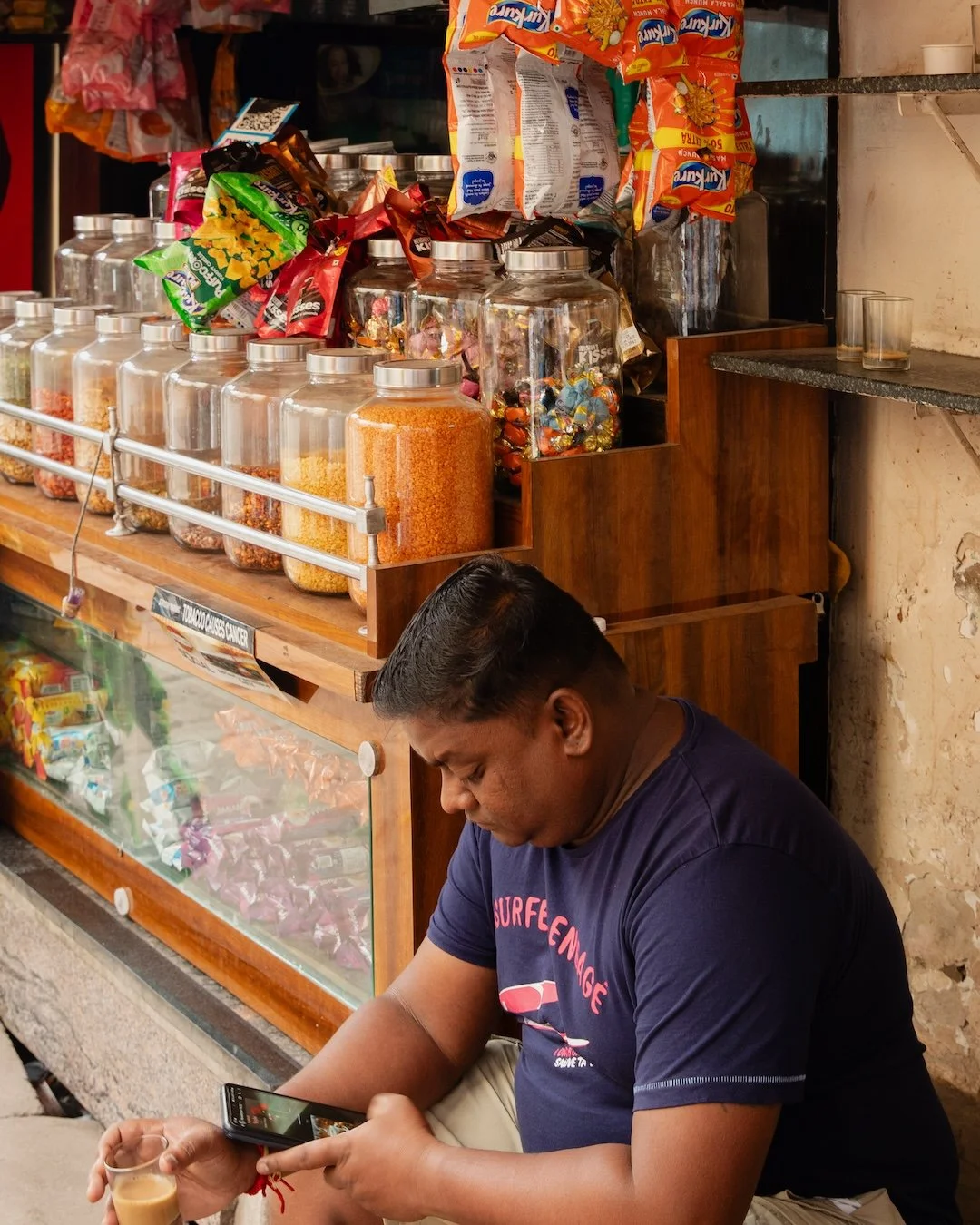 A man sitting outside a snack shop, looking at his phone while holding a small glass of coffee.