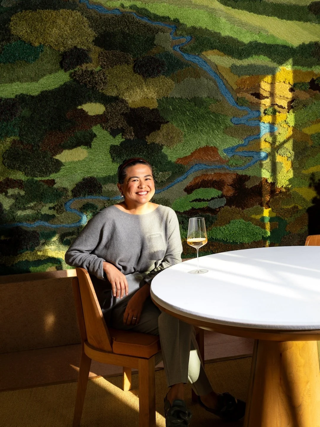 A woman smiling and sitting at a round table with a glass of white wine in a restaurant, with a colorful landscape mural of trees and a river on the wall behind her.