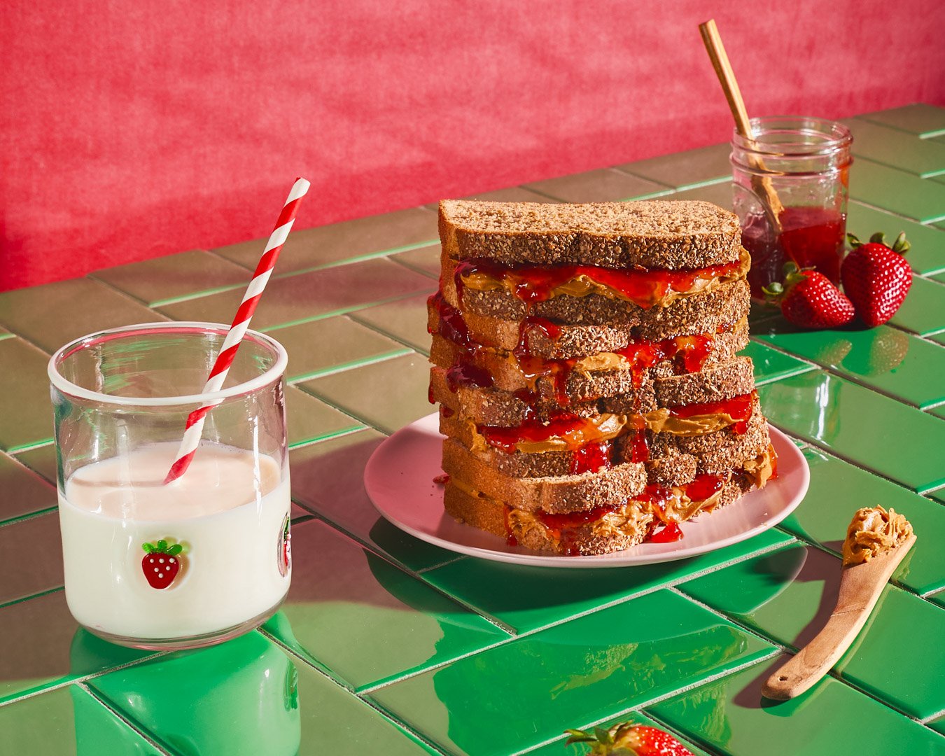 Stack of peanut butter and jelly sandwiches with visible jelly and peanut butter, on a pink plate. A glass of milk with a red and white striped straw, a jar of jelly with a spoon, and fresh strawberries on a green tiled table, with a pink wall in the