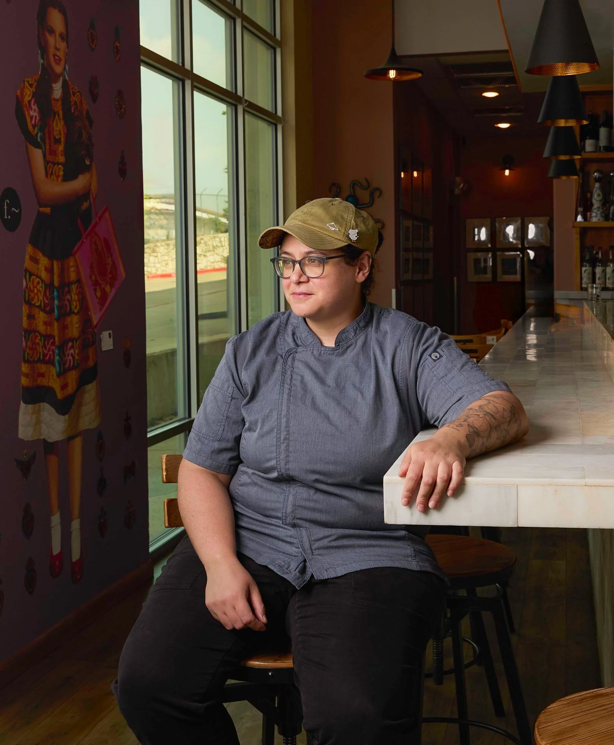 A man wearing glasses and a khaki cap with pins, sitting at a bar or cafe counter by a large window, with a brick wall and colorful artwork in the background.