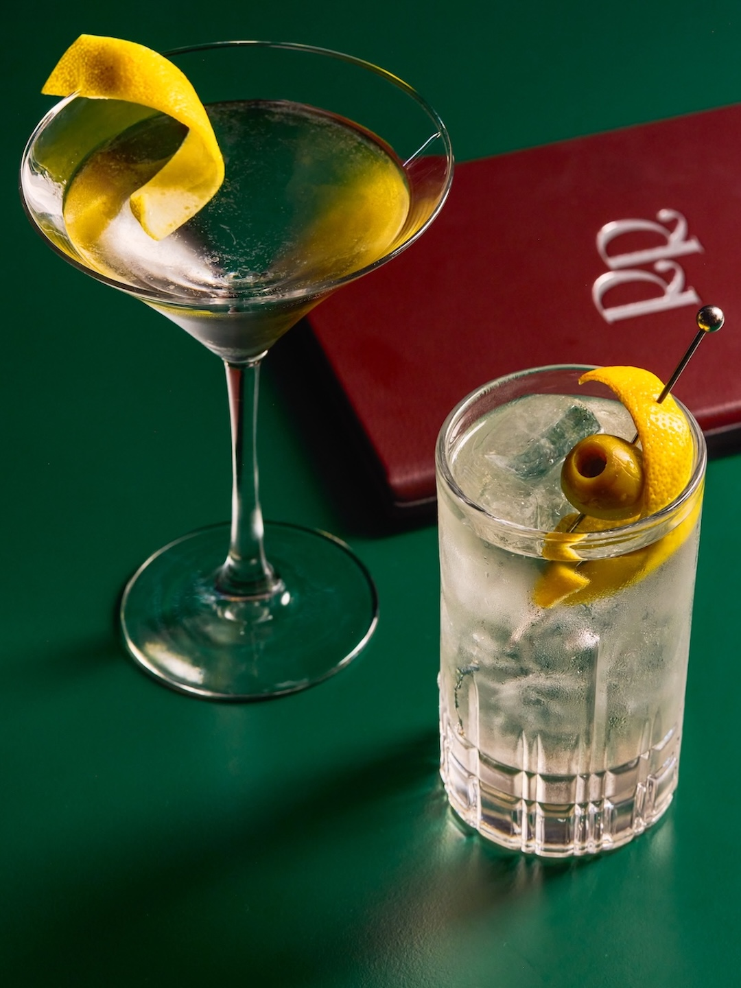 A martini glass with a lemon twist garnish and a highball glass with ice, lemon twist, and green olives, on a green surface with a red menu or book in the background.