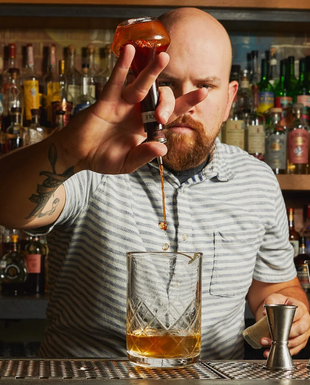Bald bartender with tattoos preparing a cocktail behind the bar, with liquor bottles in the background.