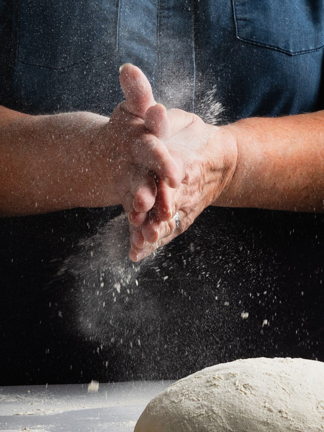 A person clapping hands together with flour dusting in the air, with a ball of dough in the foreground, wearing a dark shirt.