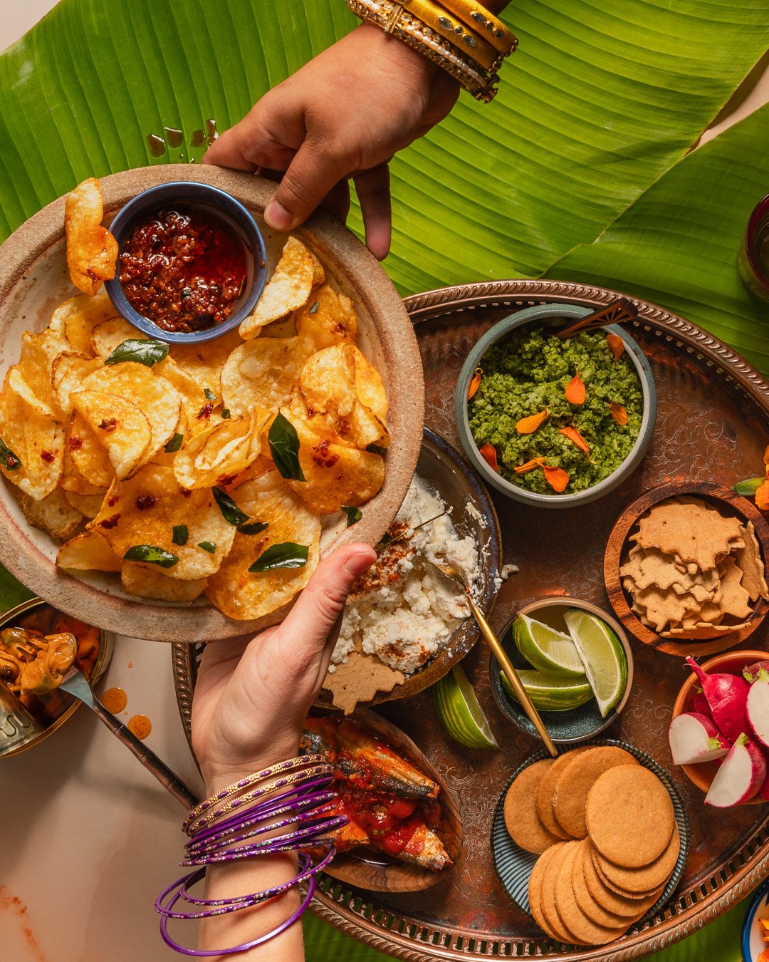 A hand holding a plate of fried chips with a small bowl of spicy salsa on top. The table is decorated with large green leaves and has various bowls containing vegetables, lime wedges, cookies, radishes, and other Indian dishes.