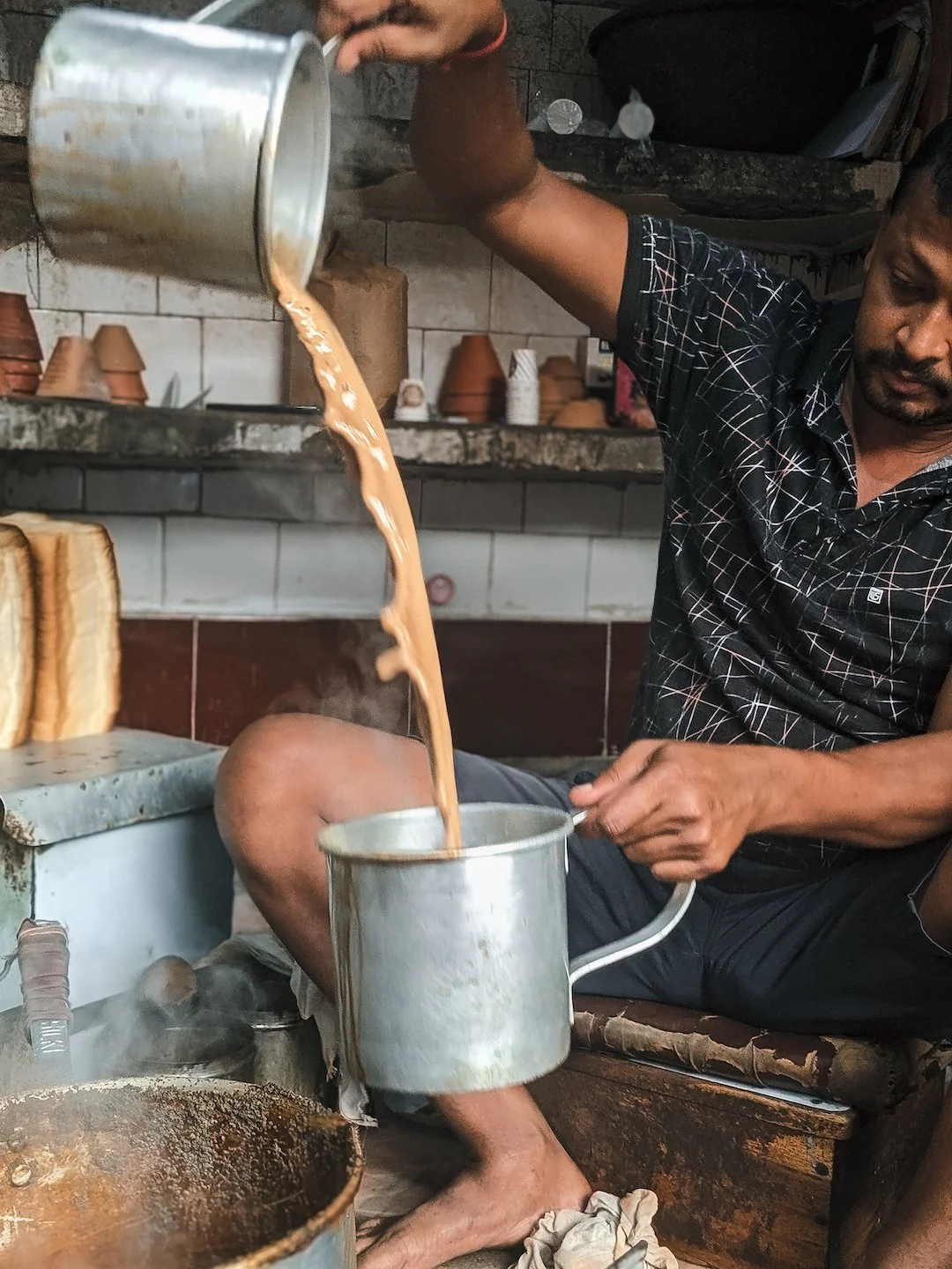 A man sitting on the floor pouring tea from a pot into a cup in a rustic kitchen with clay pots and cups in the background.